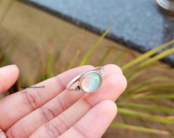 Può includere: Un anello in argento con una pietra cabochon rotonda e iridescente dalla finitura liscia e lucida. La fascia è sottile e metallica, con piccole sfere decorative in argento alla base dell'incastonatura. Il testo ShreeJaipurJewels92 è visibile.