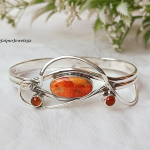 May include: A silver bracelet with an oval orange-red stone in the center and two smaller round stones on the sides. The bracelet has a unique, flowing design with the text "Shreejaipurjewels92".