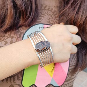 May include: Silver bangle bracelet with a brown ammonite fossil gemstone. The bracelet has five bands of silver.