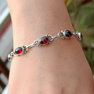 May include: A silver bracelet with five oval-shaped red gemstones. The bracelet is on a person's wrist.