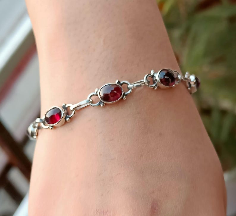 May include: A silver bracelet with five oval-shaped red gemstones. The bracelet is on a person's wrist.