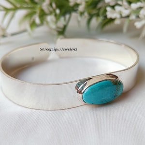 May include: A silver cuff bracelet with a large, oval turquoise stone set in the center. The bracelet is wide and has a polished finish. The background includes white flowers and greenery.