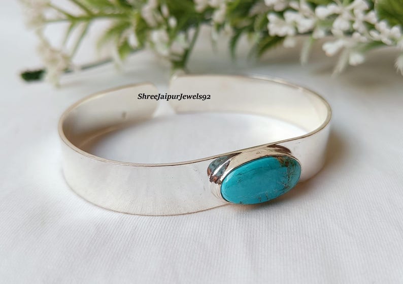 May include: A silver cuff bracelet with a large, oval turquoise stone set in the center. The bracelet is wide and has a polished finish. The background includes white flowers and greenery.