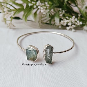 Puede incluir: Una pulsera de puño abierta plateada con dos piedras en bruto de color azul verdoso claro. La pulsera se muestra sobre una superficie blanca, con un arreglo floral en el fondo. El texto "ShreeJaipurJewels92" es visible.