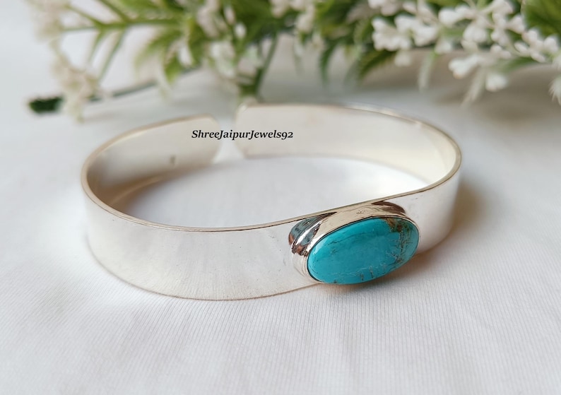 May include: A silver cuff bracelet with a large, oval turquoise stone set in the center. The bracelet is wide and has a polished finish. The background includes white flowers and greenery.