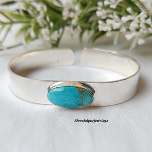 May include: A silver cuff bracelet with an oval turquoise stone set in the center. The bracelet has a wide, flat band and an open design. The background features white fabric and greenery. The text "Shreejaipurjewels92" is visible.