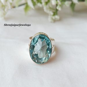 Puede incluir: Un anillo de plata con una gran gema ovalada de color azul claro. El anillo tiene una banda sencilla y un engaste que resalta las facetas de la piedra. El fondo es una tela blanca suave, con el texto "Shreejaipurjewels92" visible.