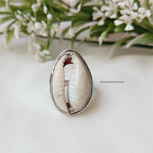 May include: A silver-toned ring featuring a natural, off-white cowrie shell. The shell is set within a smooth, oval frame. The ring is displayed on a white surface, with blurred greenery in the background. The text "Shreejaipurjewels92" is visible.