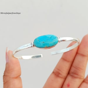 May include: A silver cuff bracelet featuring a central oval-shaped turquoise stone with natural veining. The metal band is smooth and polished with a bright metallic finish. The text ShreeJaipurJewels92 is visible in the upper left corner.
