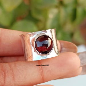 May include: A silver ring with a large, round, deep red gemstone. The ring has a wide band with a slightly curved design. The gemstone is set in a circular bezel. The text "JasperSilver2023" is visible on the ring.