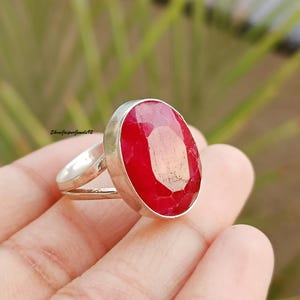 May include: A silver ring featuring a large, oval-cut, vibrant red gemstone. The ring's band splits into two delicate strands on either side of the gemstone. The background is blurred, with green and brown tones.