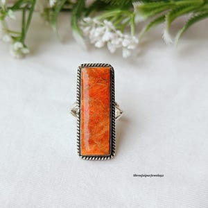 Könnte beinhalten: Ein rechteckiger Ring mit einem orangefarbenen Stein, der in einem silberfarbenen Metall gefasst ist. Der Stein hat ein strukturiertes Aussehen und ist von einem gedrehten, seilartigen Rand umgeben. Der Ring wird auf einer weißen Oberfläche präsentiert.