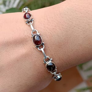 May include: A silver bracelet with five oval-shaped red gemstones. The bracelet is on a person's wrist.