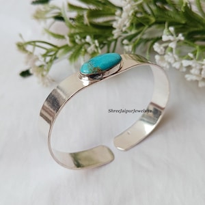 May include: A silver cuff bracelet with a turquoise stone. The open-ended bracelet has a polished finish and a rectangular band. The turquoise stone is oval-shaped and set in a silver bezel. The background includes white flowers and greenery.