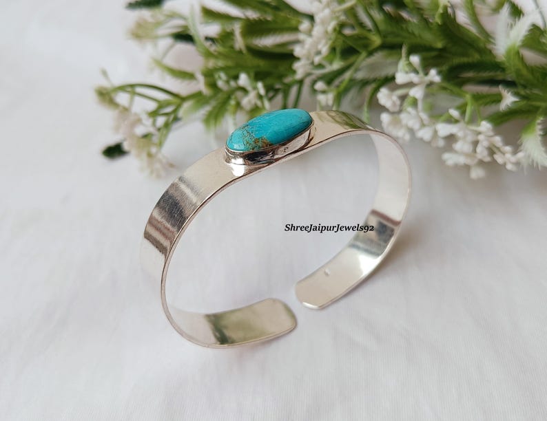 May include: A silver cuff bracelet with a turquoise stone. The open-ended bracelet has a polished finish and a rectangular band. The turquoise stone is oval-shaped and set in a silver bezel. The background includes white flowers and greenery.