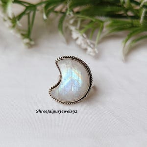 May include: A crescent-shaped ring with a silver-colored band and a large, iridescent moonstone. The moonstone displays a play of colors, including blues and greens. The ring is set against a white background with greenery in the top corners. The text "ShreeJaipurJewels92" is at the bottom.