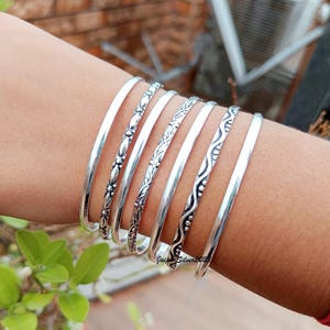 Könnte beinhalten: Ein Set aus sieben silbernen Armreifen mit verschiedenen geätzten Designs. Die Armreifen sind auf dem Handgelenk einer Person gestapelt.