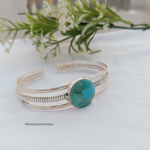May include: A silver cuff bracelet featuring a round, polished turquoise stone at the centre. The band is constructed from multiple thin silver wires with decorative coiled wire detailing along the sides. The text ShreeJaipurJewels92 is visible.