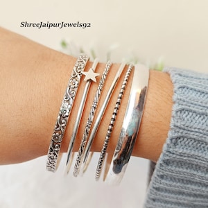 May include: A collection of silver bangles on a wrist. The bangles feature various designs, including floral patterns, a star charm, and a smooth, polished finish. The text "Shreejaipurjewels92" is visible.