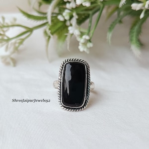 Puede incluir: Anillo rectangular de piedra negra con una banda plateada. La piedra preciosa está engastada en un marco plateado con un borde en forma de cuerda. El anillo está sobre una superficie blanca con follaje verde y blanco en el fondo. El texto "Shreejaipurjewels92" es visible.
