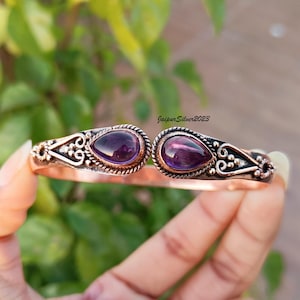Può includere: Un braccialetto rigido in rame con due pietre preziose di ametista incastonate in argento. Il braccialetto ha un design delicato e decorato.