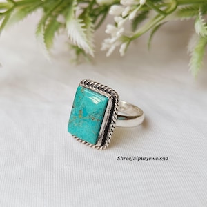 May include: A silver ring with a rectangular turquoise stone. The stone is a vibrant teal color with brown veining. The ring has a twisted rope design around the stone and a smooth, polished band. The text "Shreejaipurjewels92" is visible.