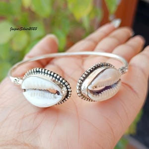 Puede incluir: Una pulsera de puño plateada con dos adornos de conchas blancas. Las conchas están engarzadas en un marco de cuentas. La pulsera es abierta y ajustable. El texto "JaipurSilver2023" es visible en el fondo.