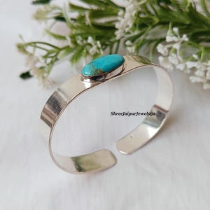 May include: A silver cuff bracelet with a turquoise stone set in the center. The bracelet has a wide, flat band and an open design. The background includes white flowers and greenery. The text "Shreejaipurjewels92" is visible on the bracelet.