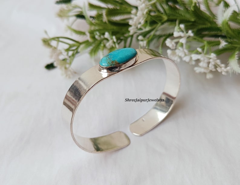 May include: A silver cuff bracelet with a turquoise stone set in the center. The bracelet has a wide, flat band and an open design. The background includes white flowers and greenery. The text "Shreejaipurjewels92" is visible on the bracelet.