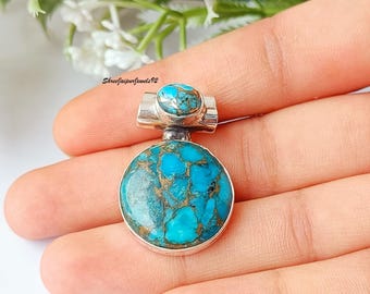 May include: A silver pendant with a large, round turquoise stone and a smaller turquoise stone on top. The turquoise has a blue-green color with brown veining. The pendant is held in a hand, with the text "ShreeJaipurJewels92" visible.