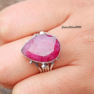 May include: A silver ring with a large, faceted, pink ruby gemstone. The ring has a delicate, intricate design with multiple bands and small silver beads.