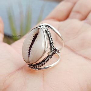 Op de afbeelding: Een zilveren ring met een kaurischelp als middelpunt. De schelp is crèmewit van kleur met een donker interieur. De ring heeft een gevlochten touwontwerp rond de schelp en een gesplitste band.