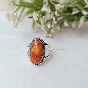 May include: Oval-shaped ring with a silver band and a large, orange-red stone. The stone has a unique, textured appearance. The ring is set against a white background with a blurred floral arrangement in the background.