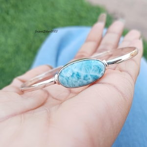 May include: A silver bangle bracelet featuring a light blue oval-shaped gemstone. The gemstone has a swirling pattern of white and blue.