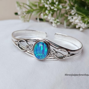Peut inclure: Bracelet manchette en argent orné d'une pierre d'opale bleue irisée ovale. Le bracelet présente des accents décoratifs en forme d'infini. Fond blanc avec de la verdure.