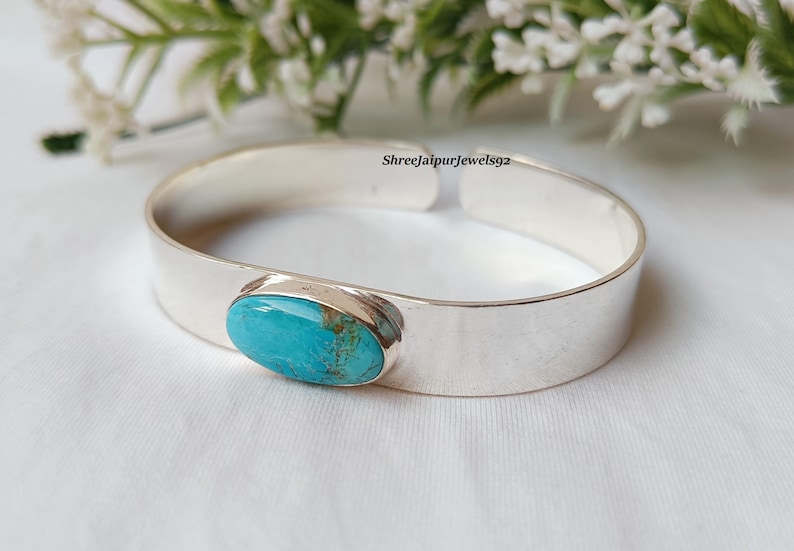 May include: A silver cuff bracelet with an oval turquoise stone. The turquoise stone has a blue-green color with brown matrix. The bracelet is on a white surface with white flowers in the background.