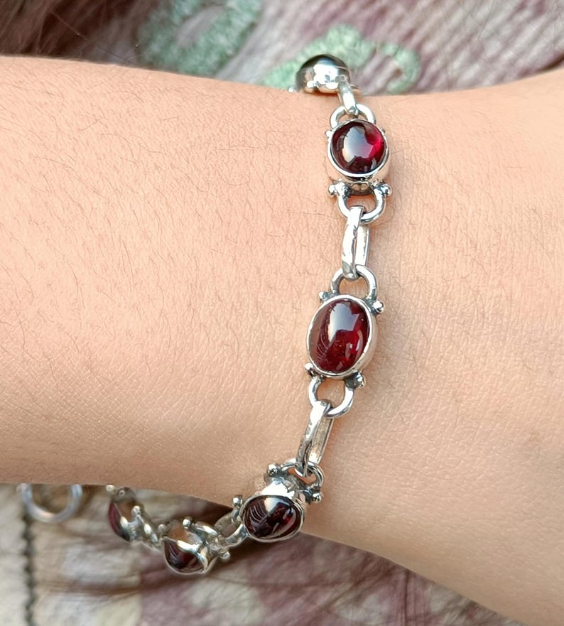 May include: A silver bracelet with red gemstone accents. The bracelet has a chain link design with oval-shaped gemstones.