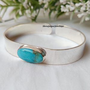 May include: A silver cuff bracelet with an oval turquoise stone. The turquoise stone is a vibrant blue-green color with brown matrix patterns. The bracelet is displayed on a white surface with greenery in the background.
