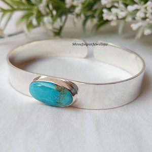 May include: A silver cuff bracelet with an oval turquoise stone. The turquoise stone has a blue-green color with brown matrix. The bracelet is on a white surface with white flowers in the background.