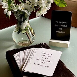 May include: A stack of white affirmation cards with black text on a brown leather-bound journal. The text on the top card reads "God will give you peace at all times and in every way". A black sign with white text reads "My prayers are powerful and effective".