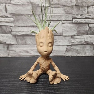 May include: A terracotta-colored Groot planter with a small air plant growing out of the top. The planter is shaped like the character Groot from the Guardians of the Galaxy movie franchise. The planter is sitting on a dark wooden surface.
