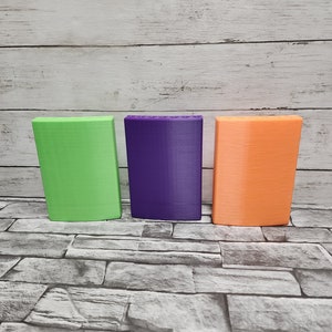 May include: Three rectangular boxes, one green, one purple, and one orange, are lined up on a gray stone surface.