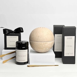 Könnte beinhalten: Ein Set mit Raumdüften in schwarzer und beiger Verpackung. Das Set enthält einen runden Diffusor, zwei kleine Flaschen mit Raumduft und zwei Schachteln mit Räucherstäbchen. Der Diffusor ist aus beigefarbenem Stein und hat einen Deckel. Die Flaschen sind schwarz und haben Etiketten mit den Aufschriften "Sand", "Revive" und "Coal". Die Räucherstäbchen befinden sich in schwarzen Schachteln mit den Aufschriften "Coal".