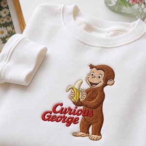 May include: A white crewneck sweatshirt featuring an embroidered graphic of the brown monkey Curious George holding a yellow banana. The text Curious George is embroidered in red script lettering below the character on the front of the garment.