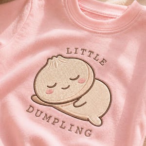 以下が含まれることがあります： 目を閉じてピンクの頬をした、眠っている餃子のキャラクターの刺繍が施されたライトピンクのトレーナー。イラストの上下には、ダークブラウンのフォントで「LITTLE DUMPLING」という文字が刺繍されています。