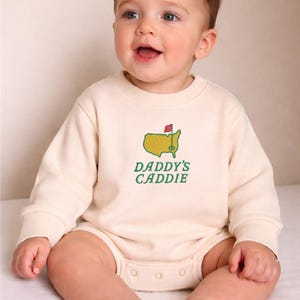 May include: A cream-colored baby onesie with long sleeves. The onesie features an embroidered design of a golf course map with a red flag and the text "DADDY'S CADDIE" in green. The baby is sitting and smiling.