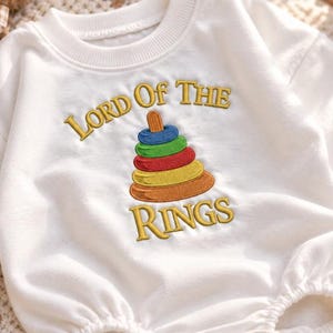May include: A white long-sleeve sweatshirt featuring an embroidered graphic of a colorful stacking ring toy. The text "LORD OF THE RINGS" is stitched in yellow thread above and below the toy, which consists of blue, green, red, yellow, and orange rings.