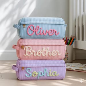 May include: Three corduroy pencil cases stacked. The top is light blue with "Oliver" in pink. The middle is pink with "Brother" in white. The bottom is lavender with "Sophia" in yellow and blue. Each case has a zip and a small handle.