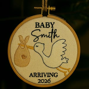 Pode incluir: Arte em bastidor bordado com uma cegonha carregando um embrulho de bebê. O texto diz "BABY Smith ARRIVING 2026". O design está em tecido creme, em um bastidor de madeira, e suspenso por barbante natural.