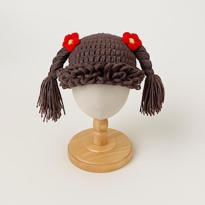 May include: A brown crochet hat with attached braids and red flower accents. The hat features a ruffled brim and is displayed on a mannequin head. The braids hang down on either side of the head, adding a playful touch to the design.